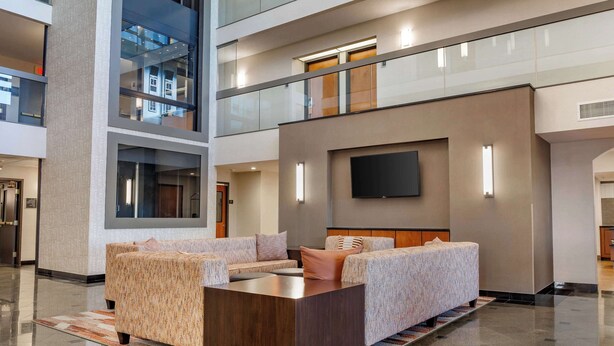 A modern hotel lobby with a high ceiling, a central seating area with sofas and a coffee table, and a flat-screen TV mounted on the wall.