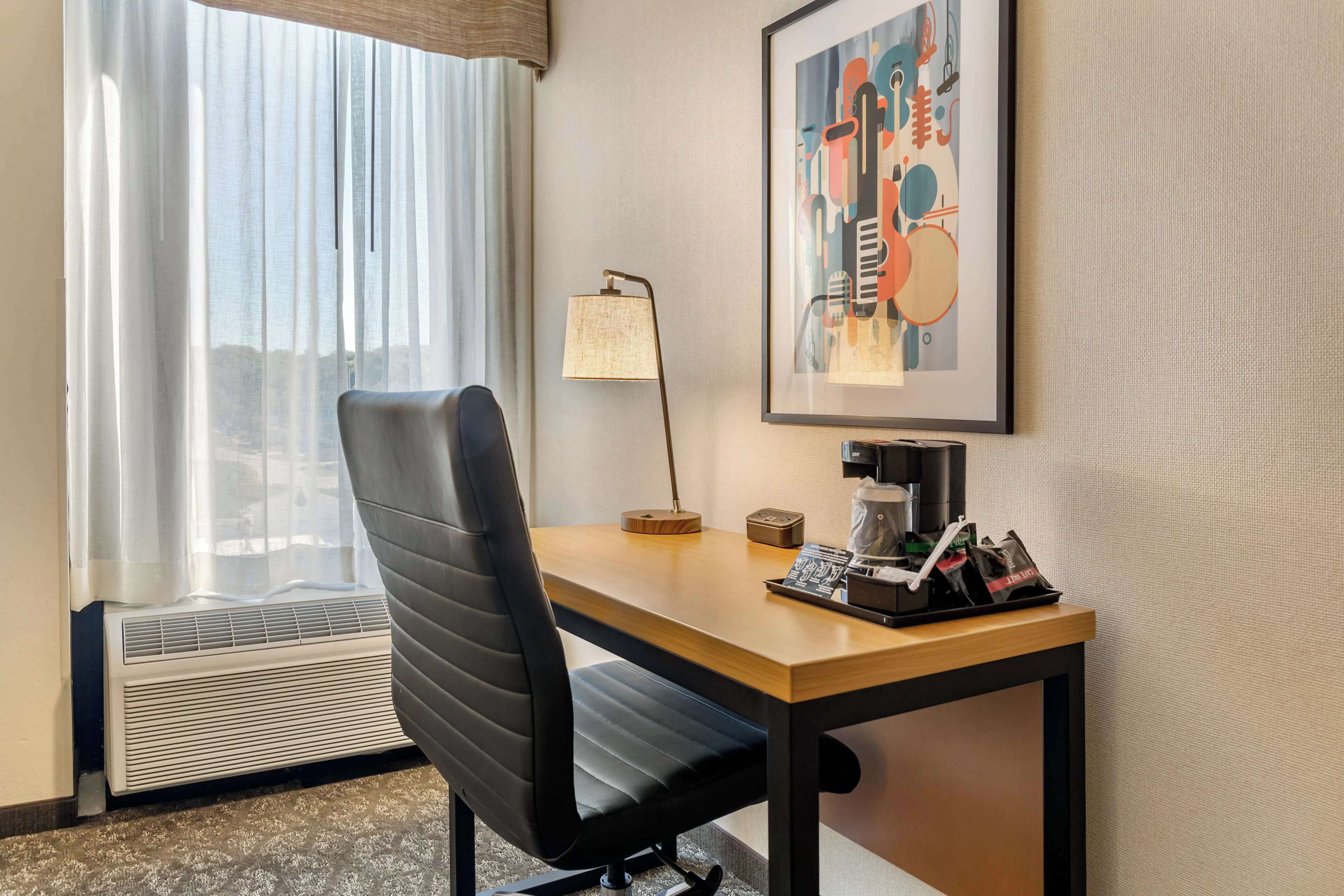 A hotel room with a desk, chair, lamp, and a framed abstract painting.