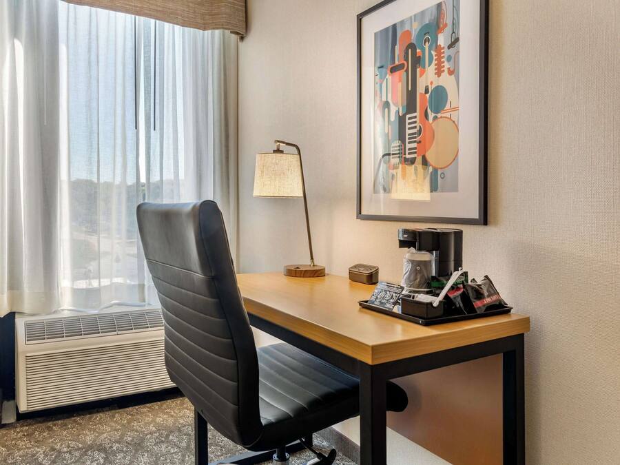 A hotel room with a desk, chair, lamp, and a framed abstract painting.