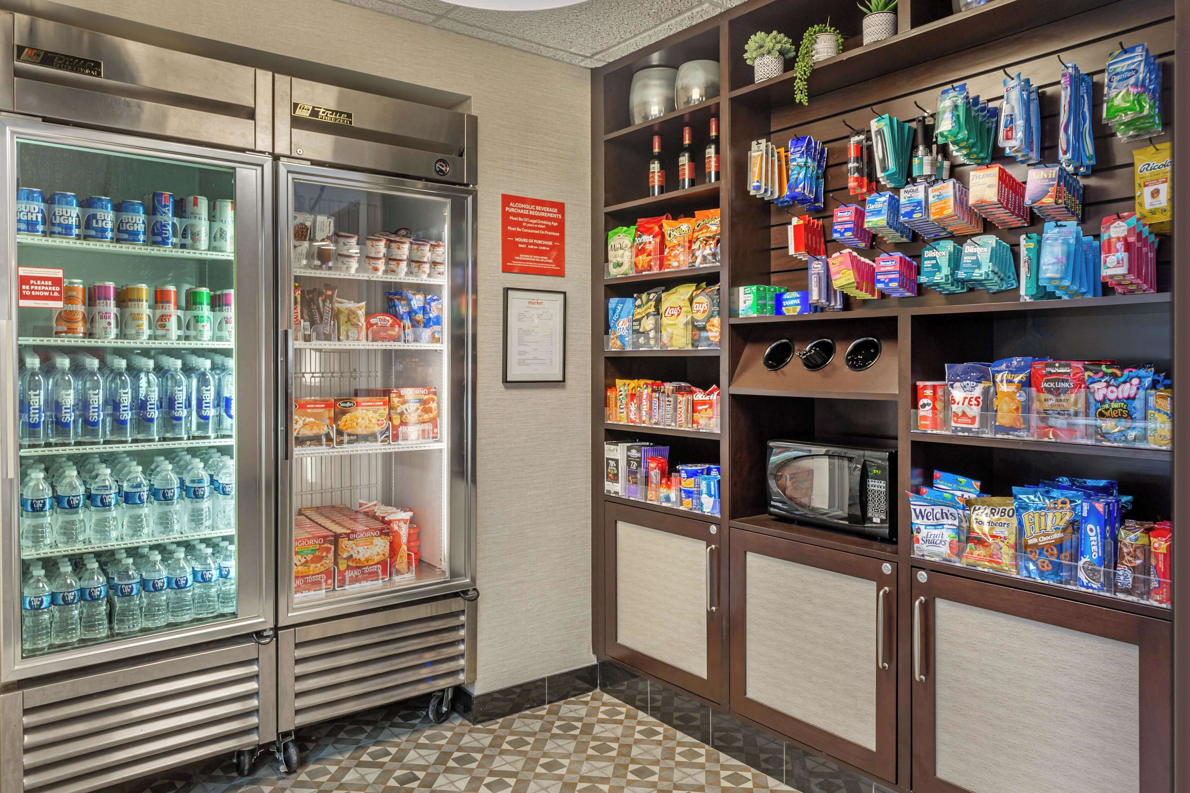 Una kitchenette con un refrigerador lleno de bebidas y aperitivos, un microondas y diferentes alimentos en los estantes.