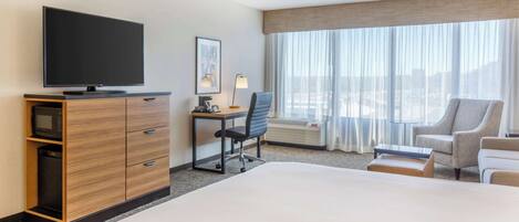 A hotel room with a large bed, a desk with a chair, a television, and a view of the city through large windows.