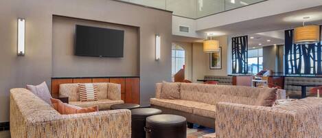 Eine geräumige Lobby mit hoher Decke, einem an der Wand befestigten Flachbildfernseher und einer Sitzecke mit Sofas und Poufs.