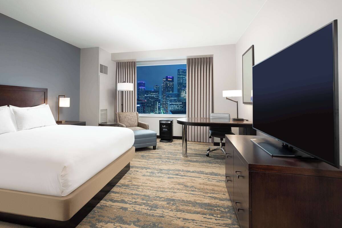 A hotel room with a large bed, a desk, a flat-screen TV, and a view of a city skyline through a window.