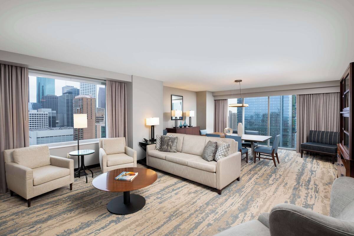 A hotel room with a living area featuring a sofa, armchairs, a coffee table, and a dining table. Large windows offer a city view.