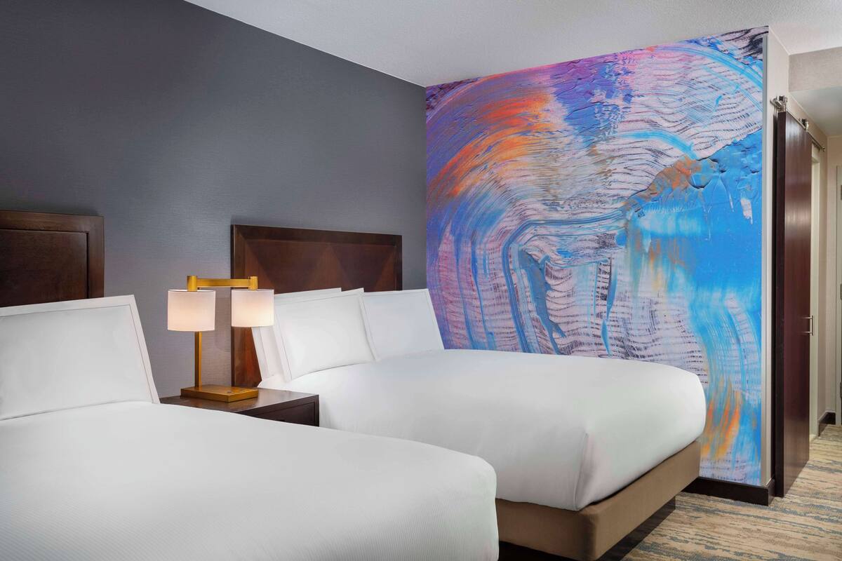 A hotel room with two beds, a large abstract painting on the wall, and a nightstand with a lamp.