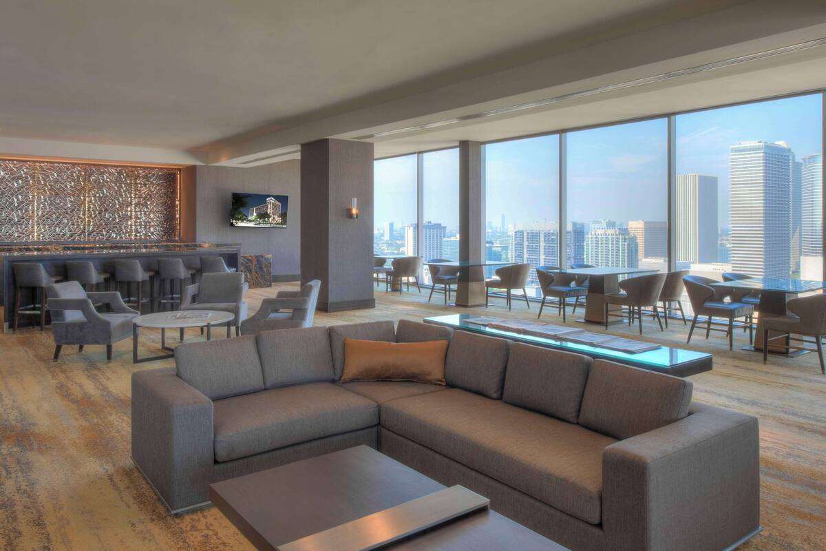 A modern hotel lobby with a large seating area, a bar, and large windows offering city views.
