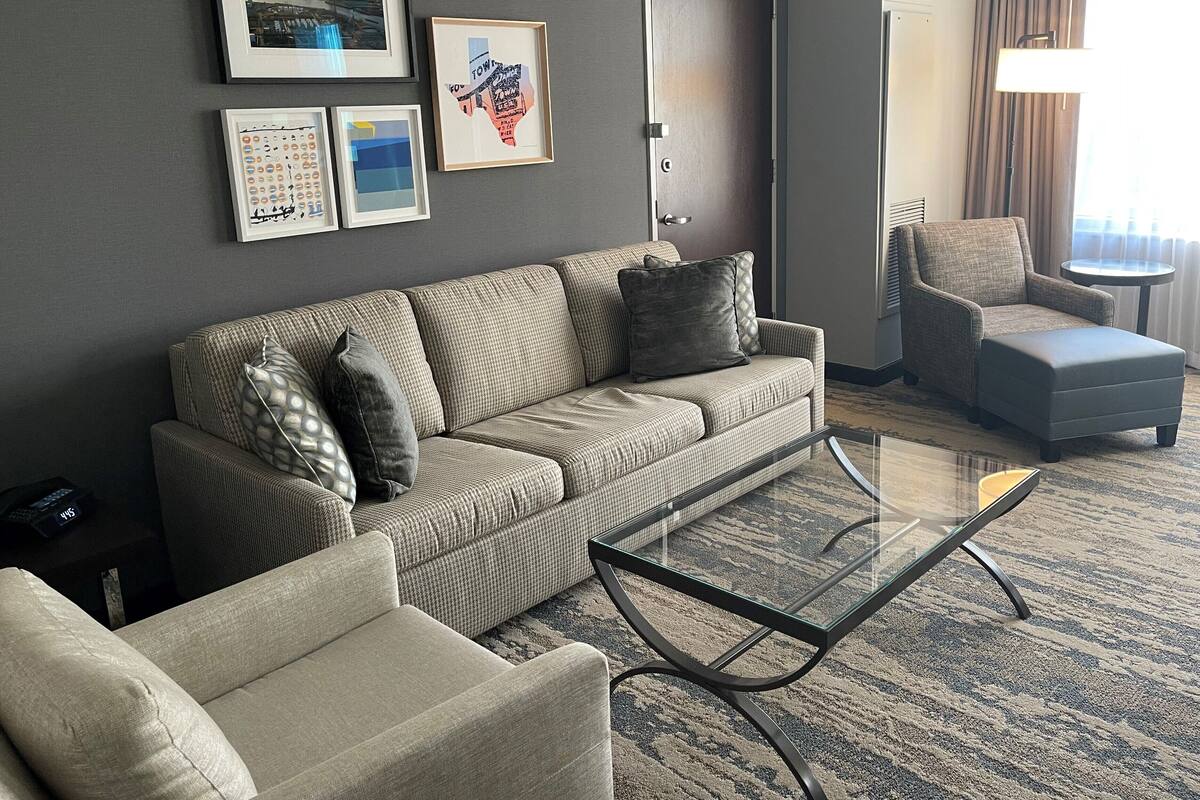 A modern hotel room with a grey sofa, a glass coffee table, a grey armchair, and a small round table. There are framed pictures on the wall and a door to the right.