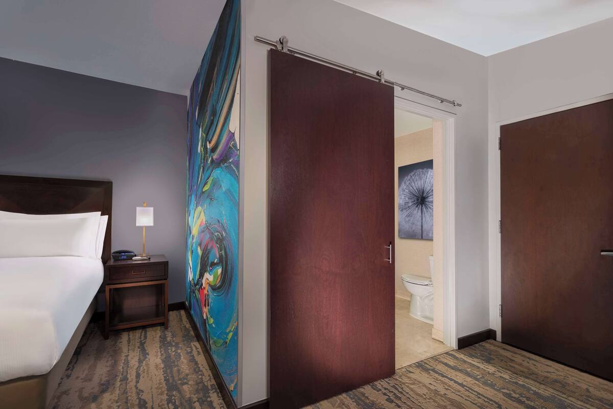 A hotel room with a bed, a nightstand, a painting, a door, and a view of the bathroom.