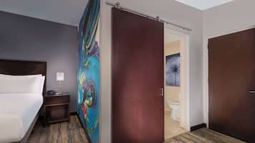 A hotel room with a bed, a nightstand, a painting, a door, and a view of the bathroom.