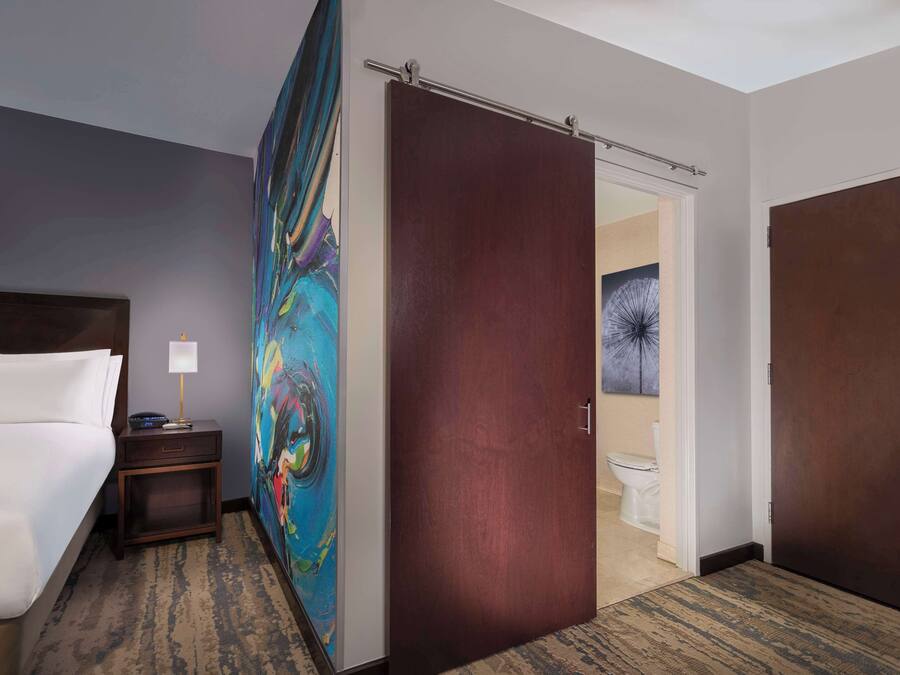 A hotel room with a bed, a nightstand, a painting, a door, and a view of the bathroom.