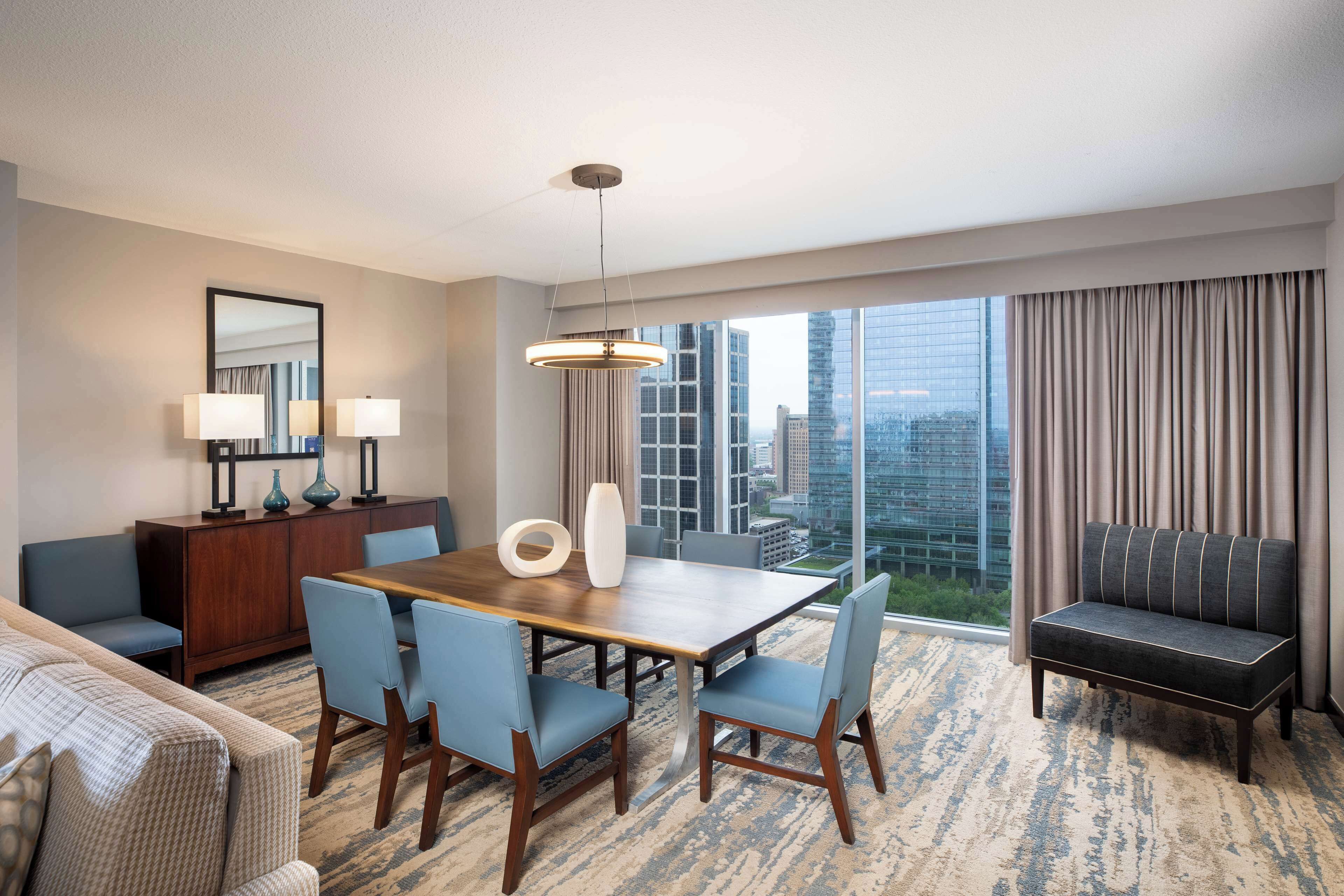 A modern hotel room with a dining area, a sofa, a large window with a city view, and a chandelier.