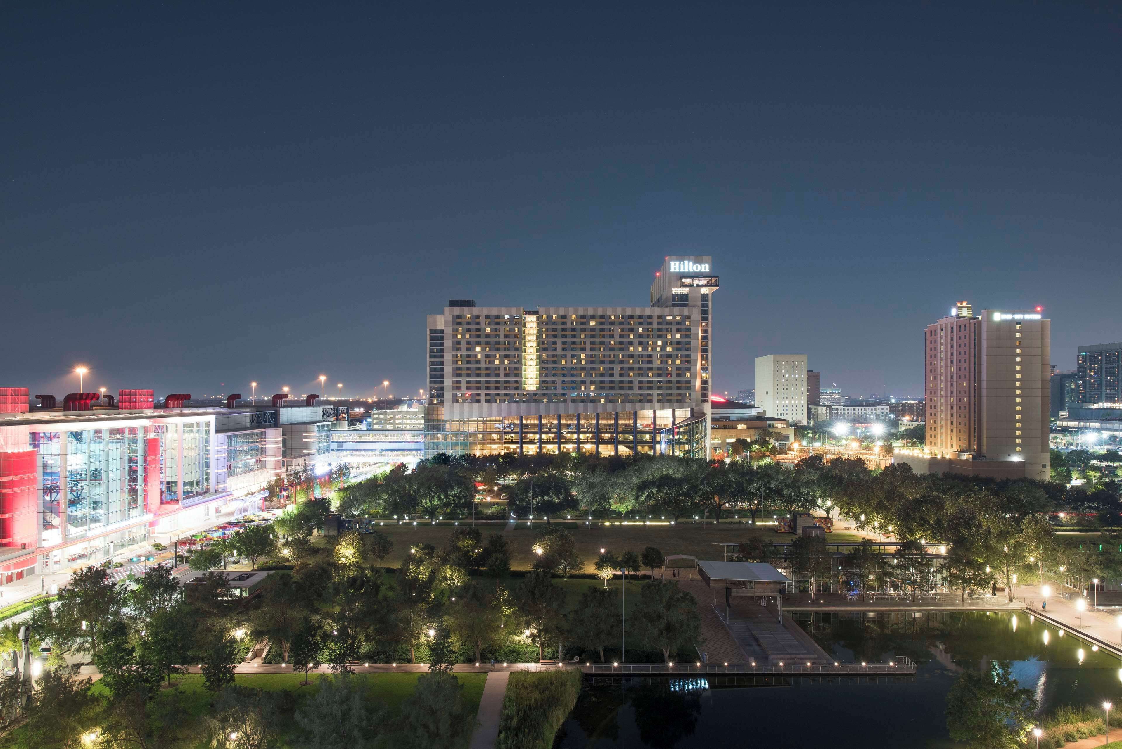 A modern hotel with a prominent Hilton sign, surrounded by lush greenery and a water feature.