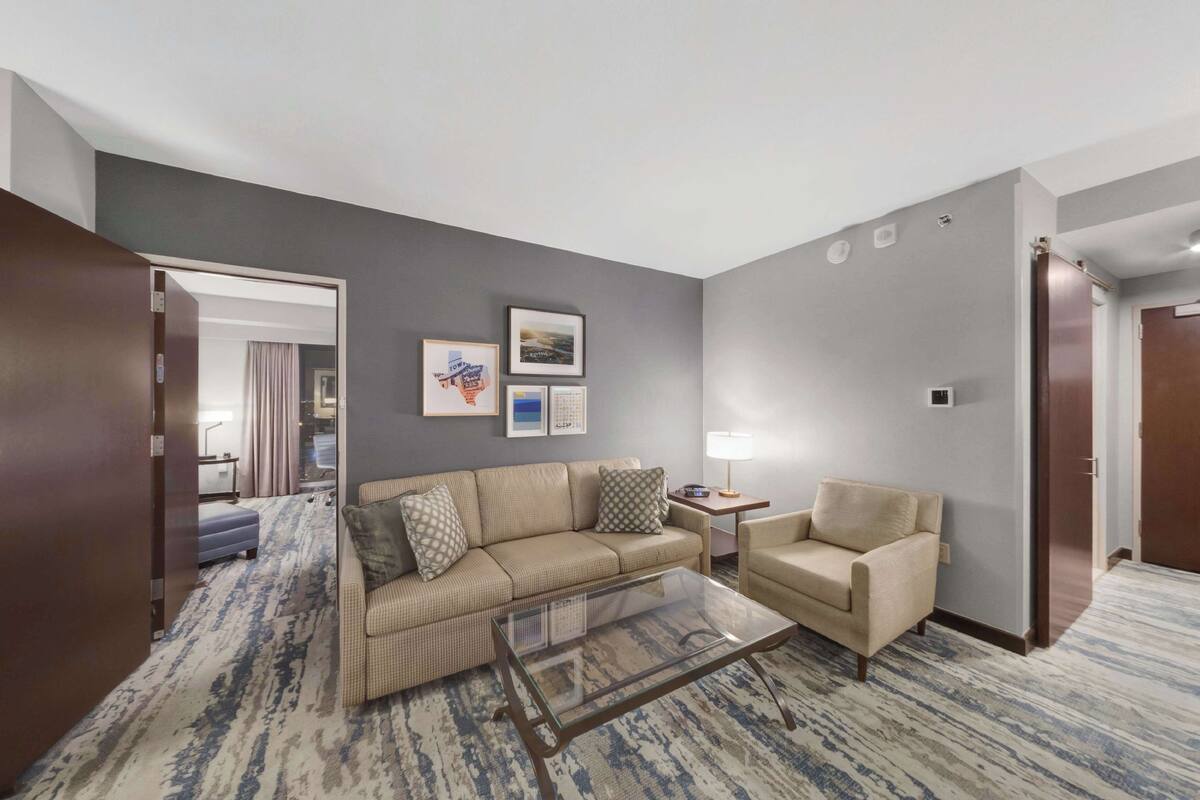 A modern hotel room with a sofa, armchair, and a glass coffee table. There are framed pictures on the wall, a lamp on a side table, and a door leading to another room.