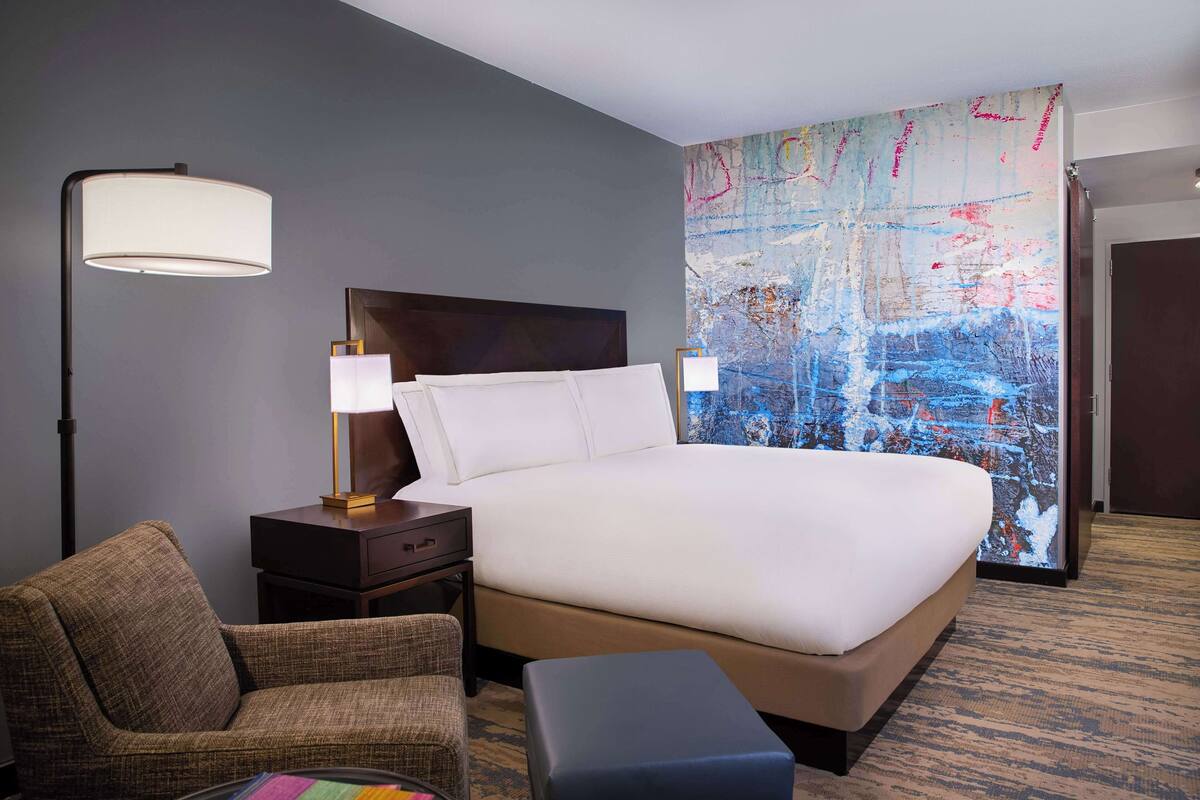 A hotel room with a large bed, a nightstand, a lamp, a chair, and a colorful abstract painting on the wall.