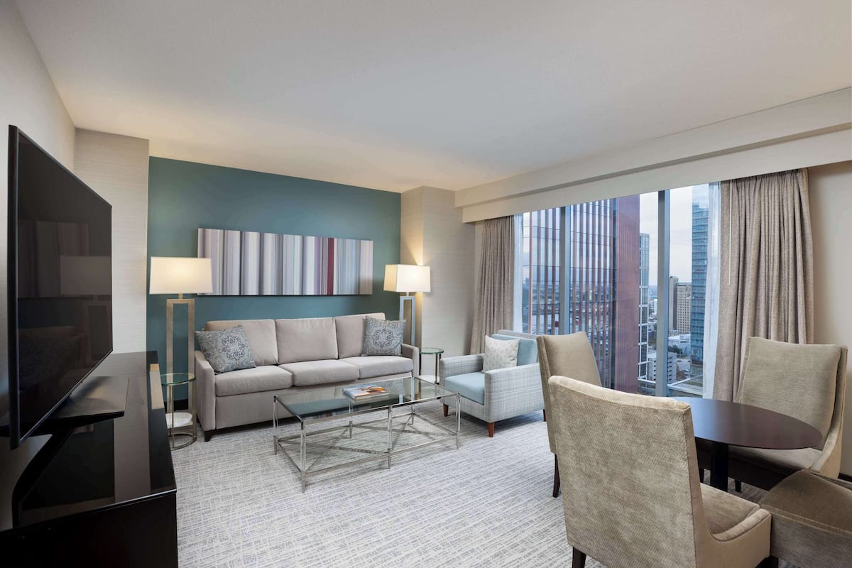 A modern hotel room with a sofa, armchairs, a coffee table, a TV, and a large window with a city view.