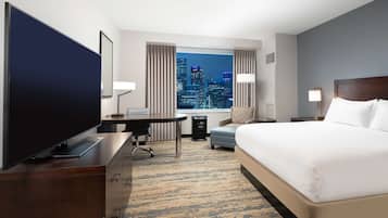 A hotel room with a large bed, a desk with a computer, a TV, and a view of a city skyline.