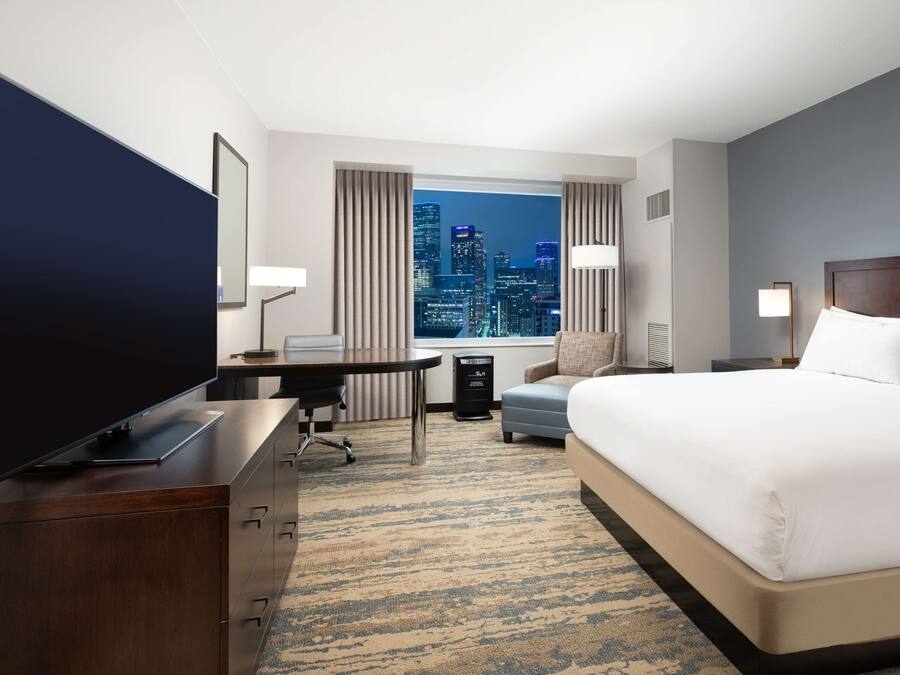 A hotel room with a large bed, a desk with a computer, a TV, and a view of a city skyline.