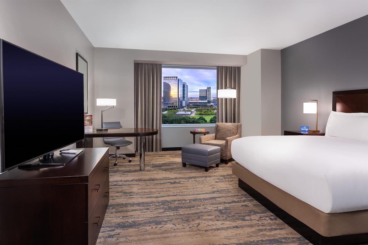 A hotel room with a large bed, a desk with a chair, a television, a small table, and a view of a cityscape.
