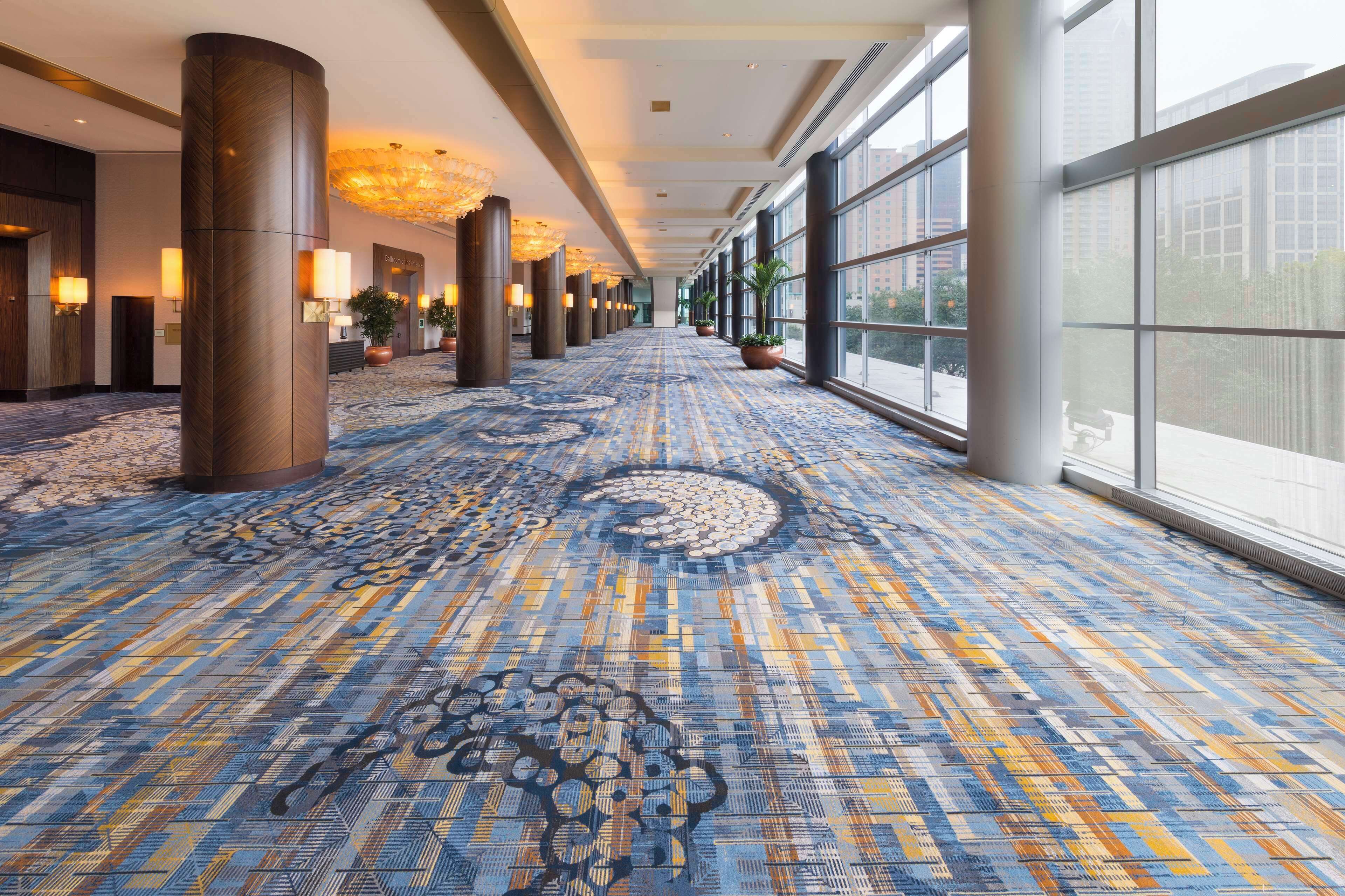 A hotel corridor with patterned carpet, large windows, and potted plants.