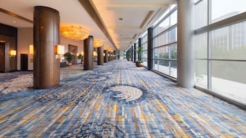 A hotel corridor with patterned carpet, large windows, and potted plants.