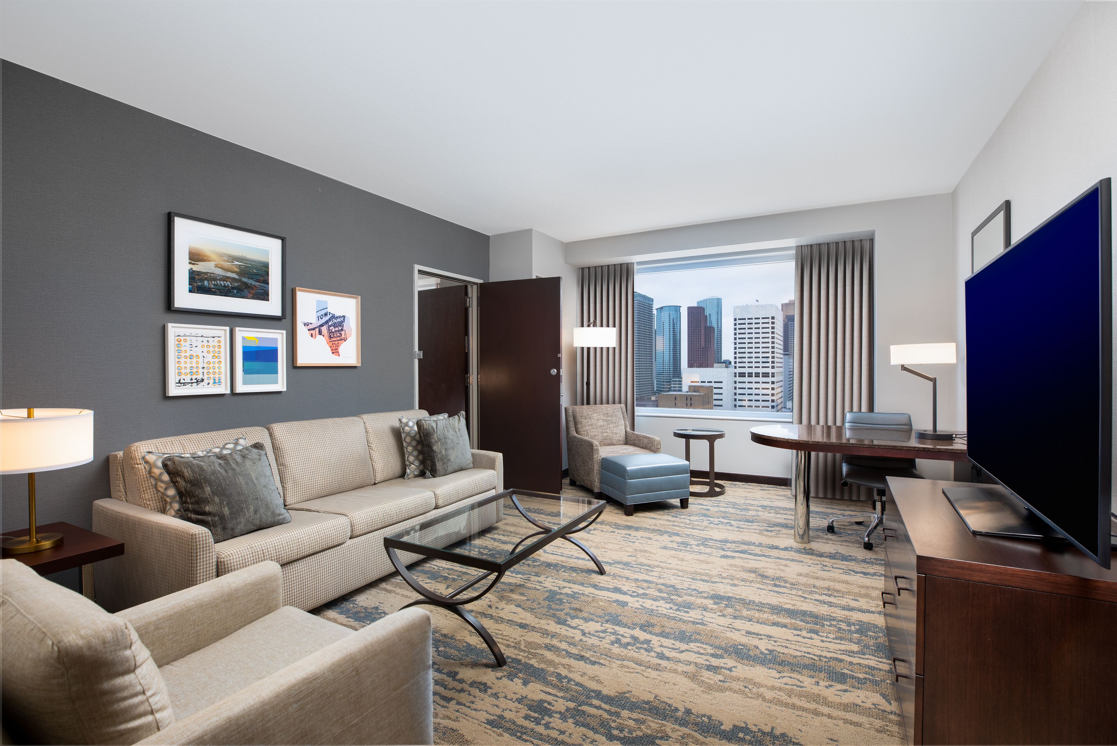 A modern hotel room with a large window, a sofa, a coffee table, a desk, and a television.