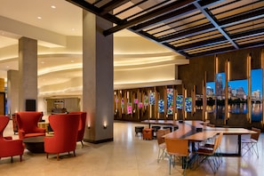 A spacious lobby with a modern design, featuring a large seating area with red chairs and a long table, a reception desk, and a cityscape view through large windows. - Hilton Austin (Austin)