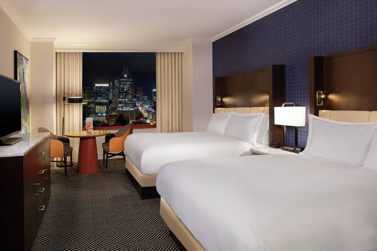 A hotel room with two beds, a desk, a chair, a TV, and a city view.