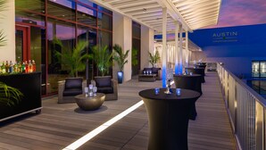 Een moderne buitenlounge met zitplaatsen en tafels, een bar met flessen, en zicht op het congrescentrum op de achtergrond.