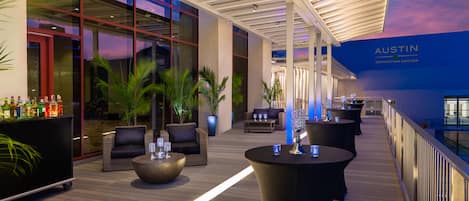 A modern outdoor lounge area with seating and tables, a bar with bottles, and a view of the convention center in the background.