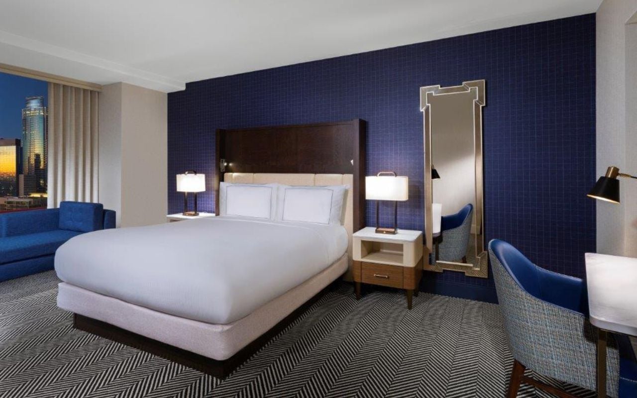A hotel room with a large bed, a nightstand, a mirror, a blue armchair, and a city view.