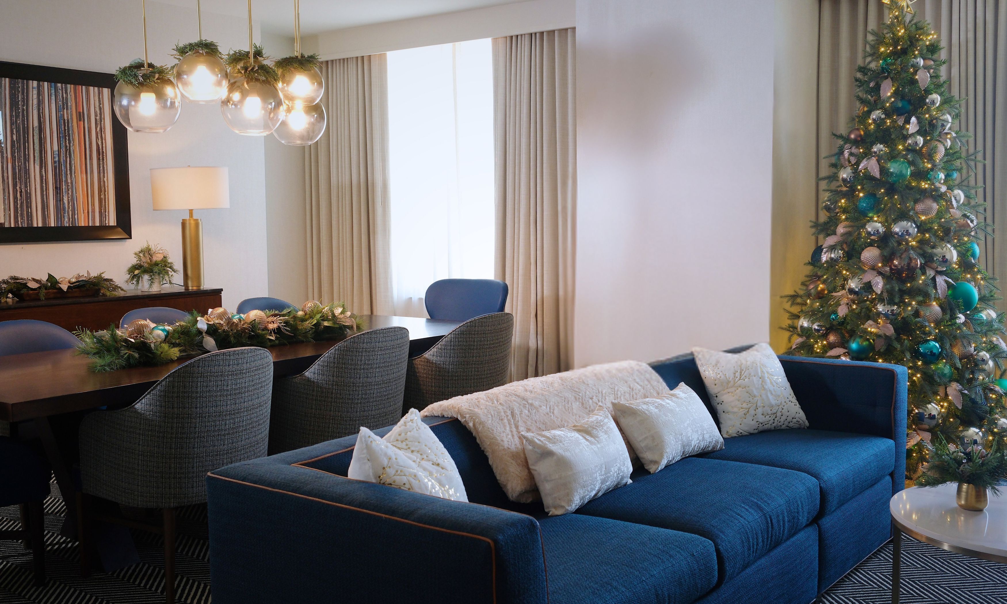 Une chambre d’hôtel moderne avec un canapé bleu, une table à manger avec des chaises, un sapin de Noël et une grande fenêtre avec des rideaux.