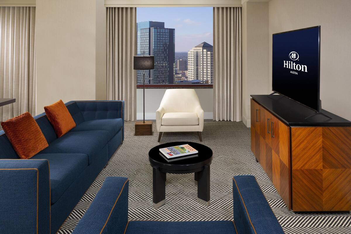 A hotel room with a sofa, armchair, coffee table, TV, and a view of city buildings.