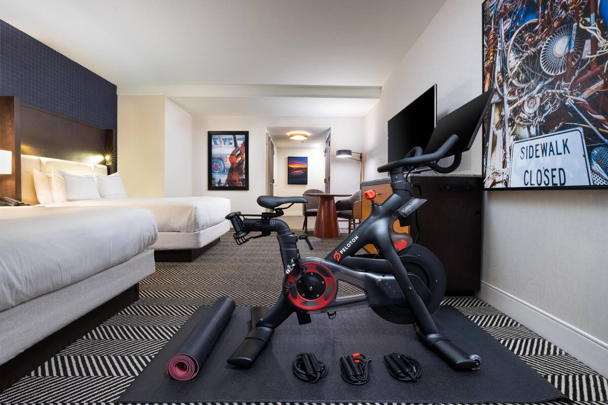A hotel room with a bed, exercise bike, and a painting of a bicycle on the wall.