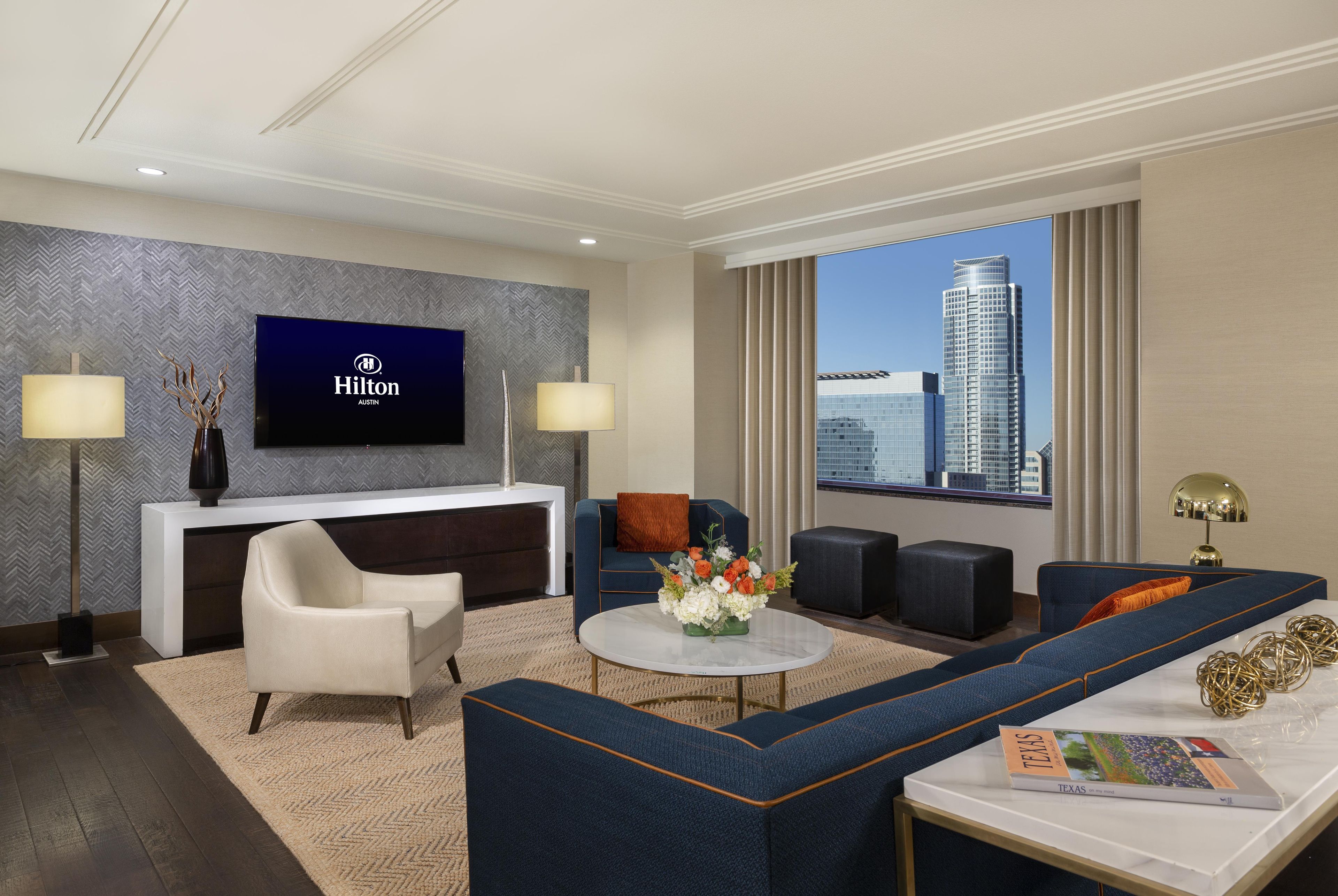 A modern hotel room with a flat-screen TV, a sofa, a coffee table, and a view of the cityscape.