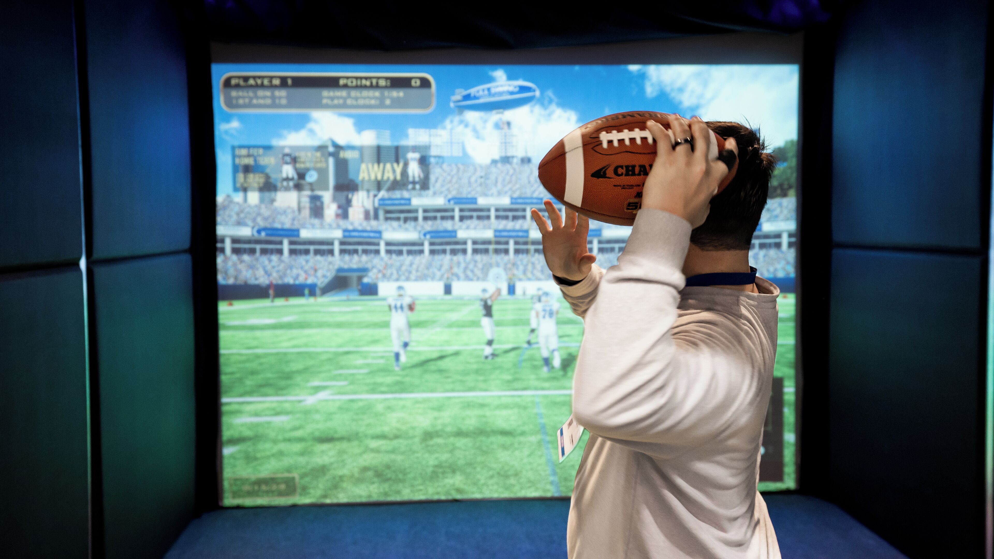A person holding a football in front of a large screen displaying a football game.