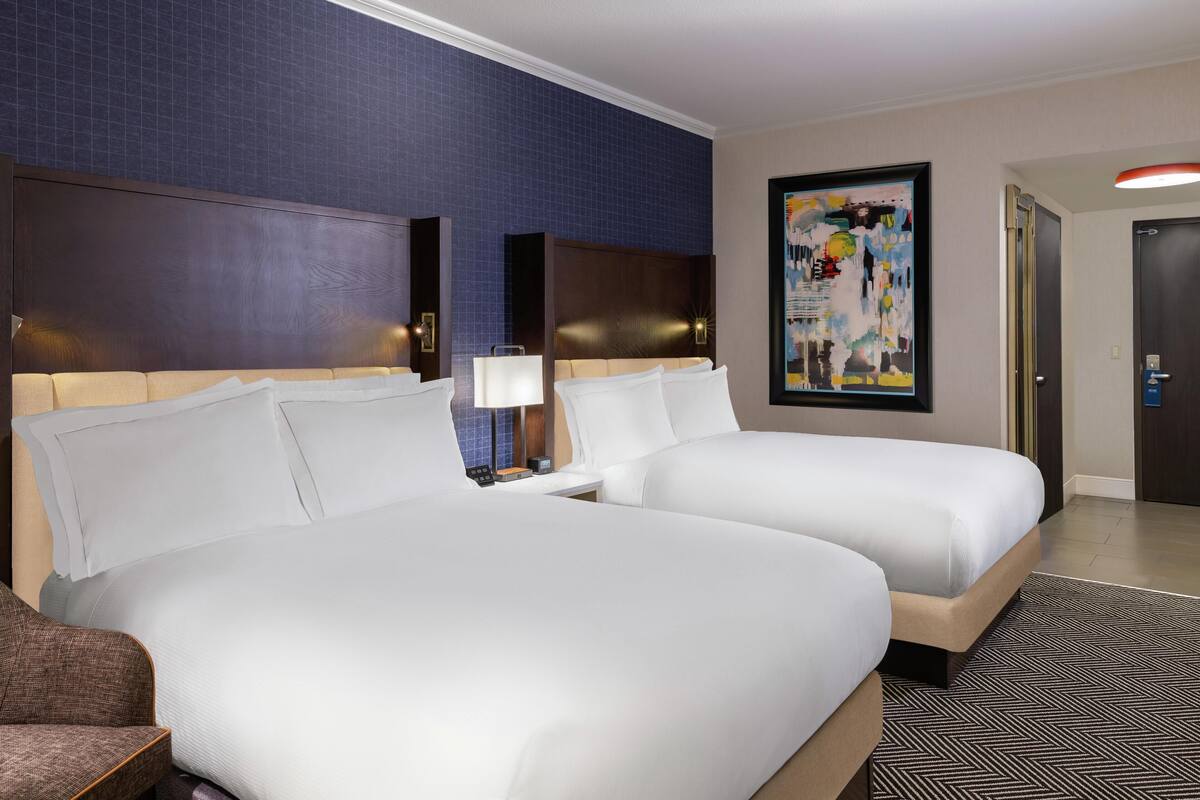 A hotel room with two beds, a nightstand, a lamp, a framed picture on the wall, and a door.