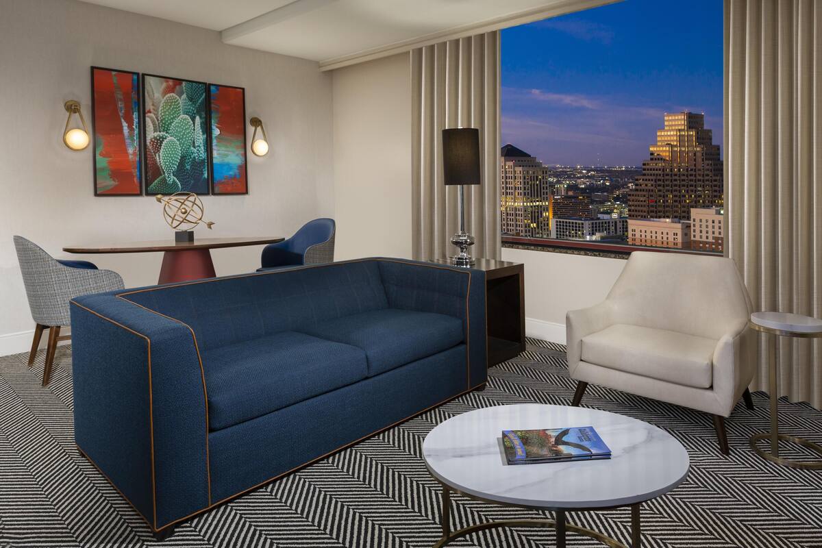 A modern hotel room with a blue sofa, a white armchair, a round coffee table, a desk with a chair, and a large window with a city view.
