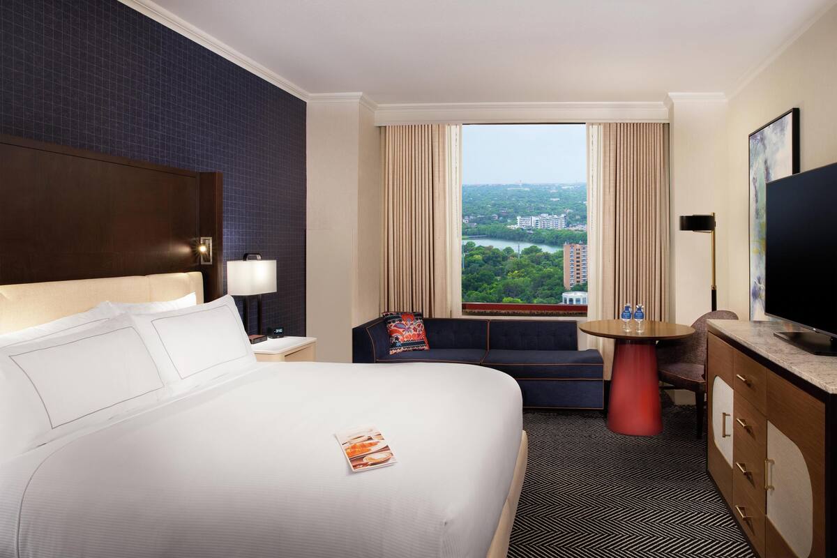 A hotel room with a large bed, a sofa, a small table, a TV, and a window with a city view.