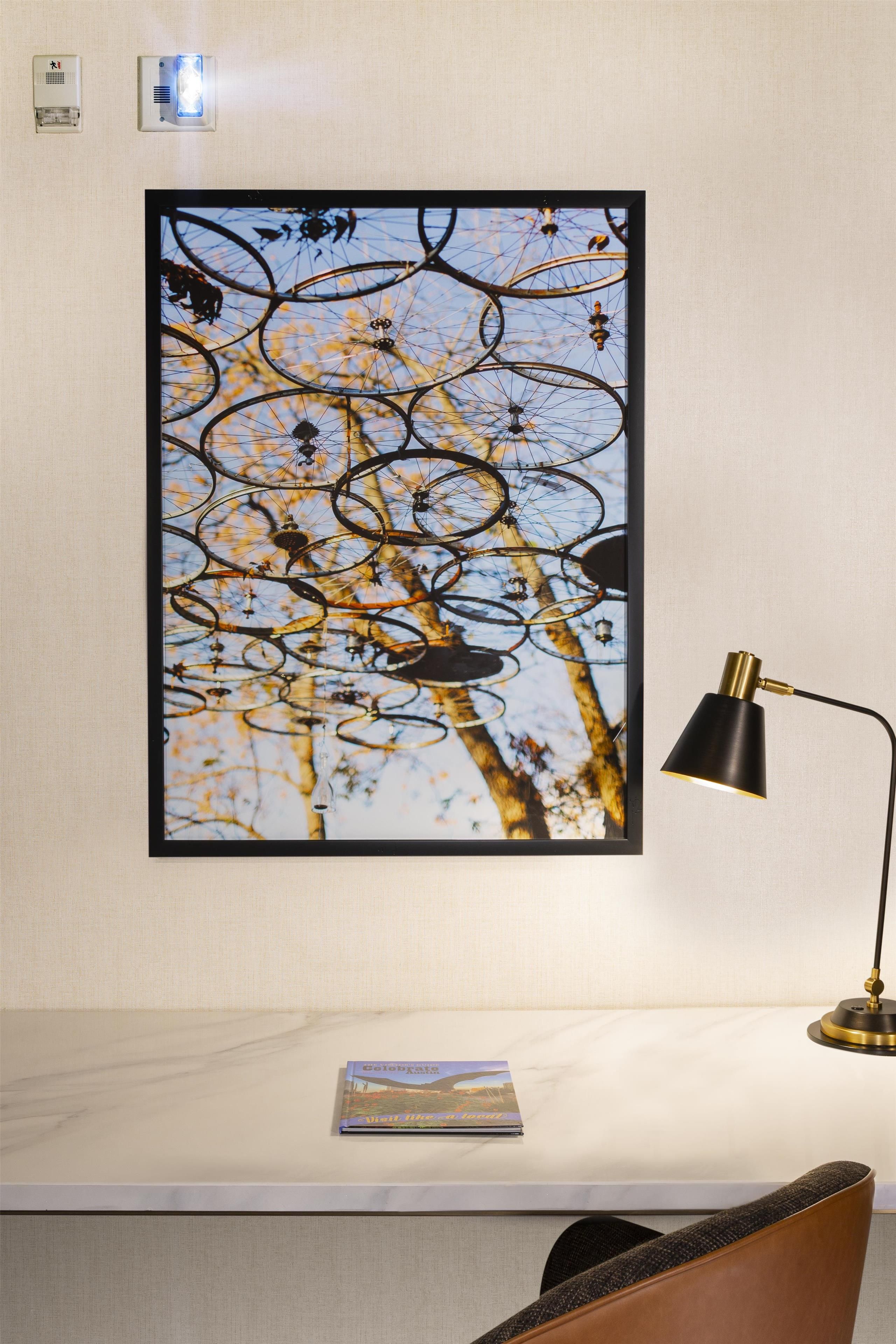A framed artwork of bicycle wheels on a wall, a desk with a lamp, and a book titled 'Celebrate Austin.'