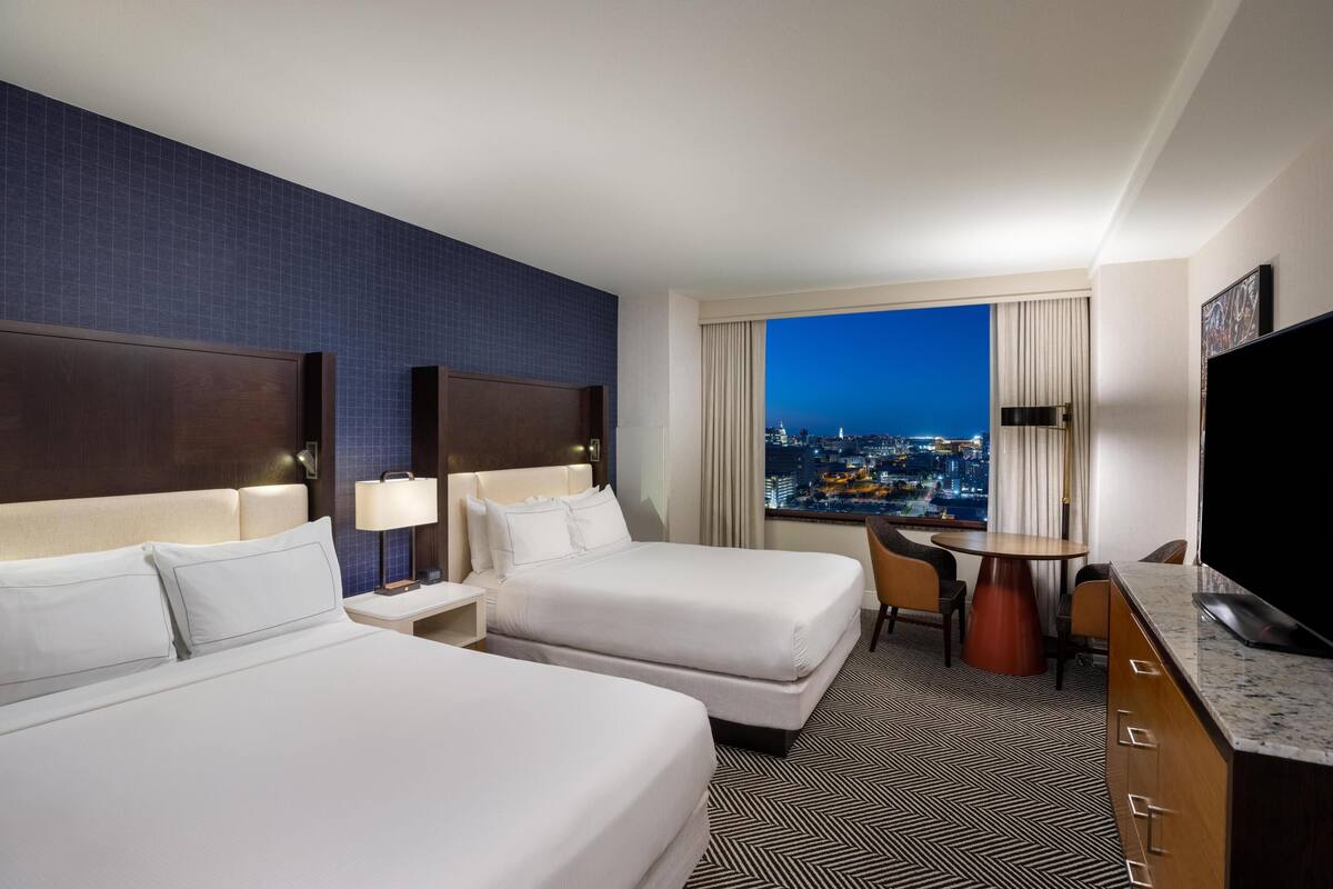 A hotel room with two beds, a TV, a desk with a chair, and a view of a cityscape at night.