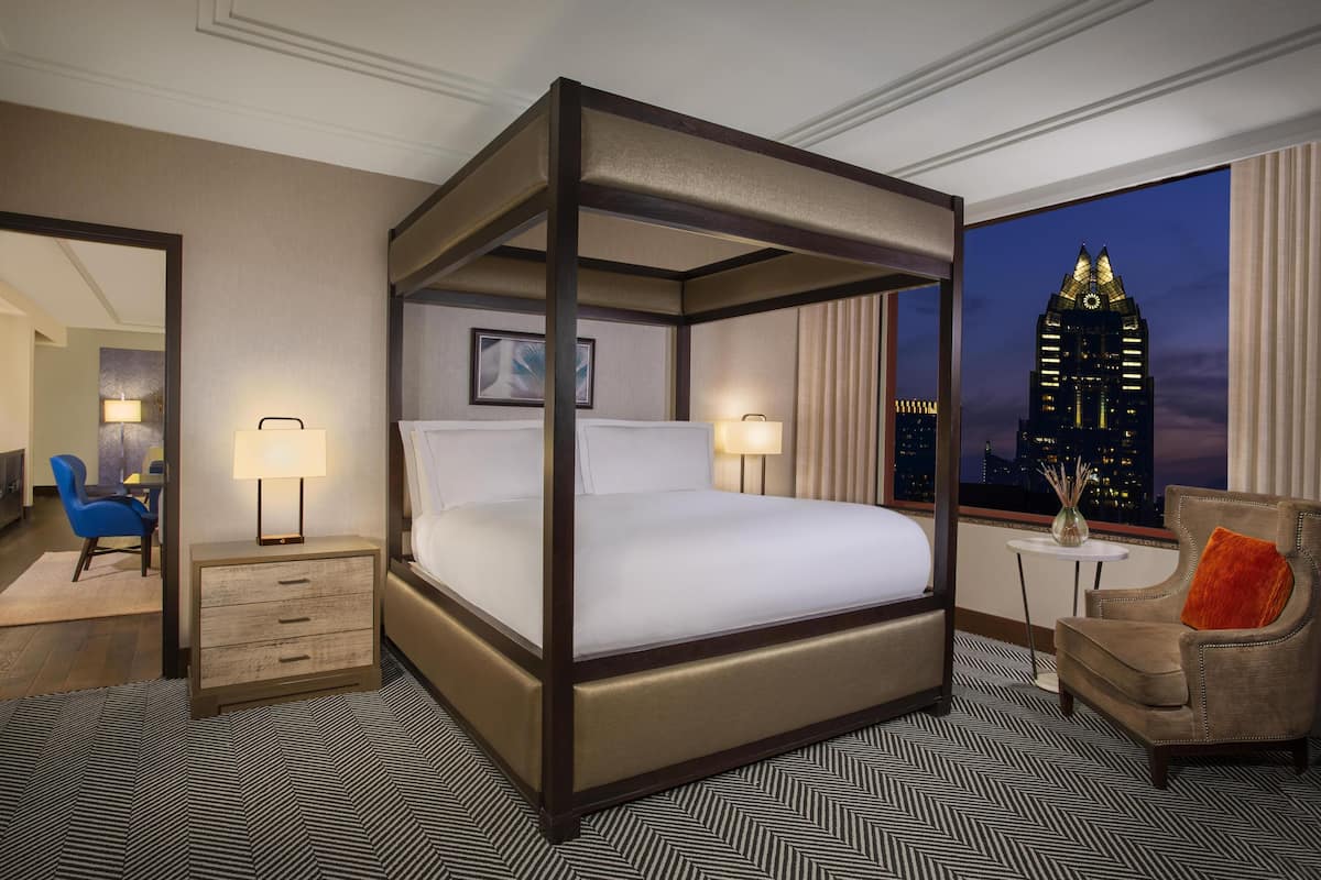 A four-poster bed with a nightstand, a chair, and a view of a cityscape at dusk.