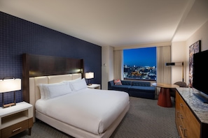 A hotel room with a bed, nightstands, a sofa, a TV, and a view of a cityscape at night.