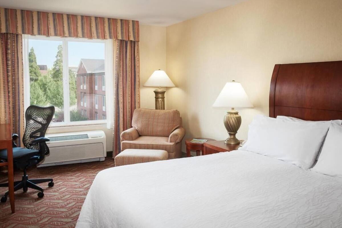 A hotel room with a bed, two chairs, a desk, a lamp, and a window with curtains.