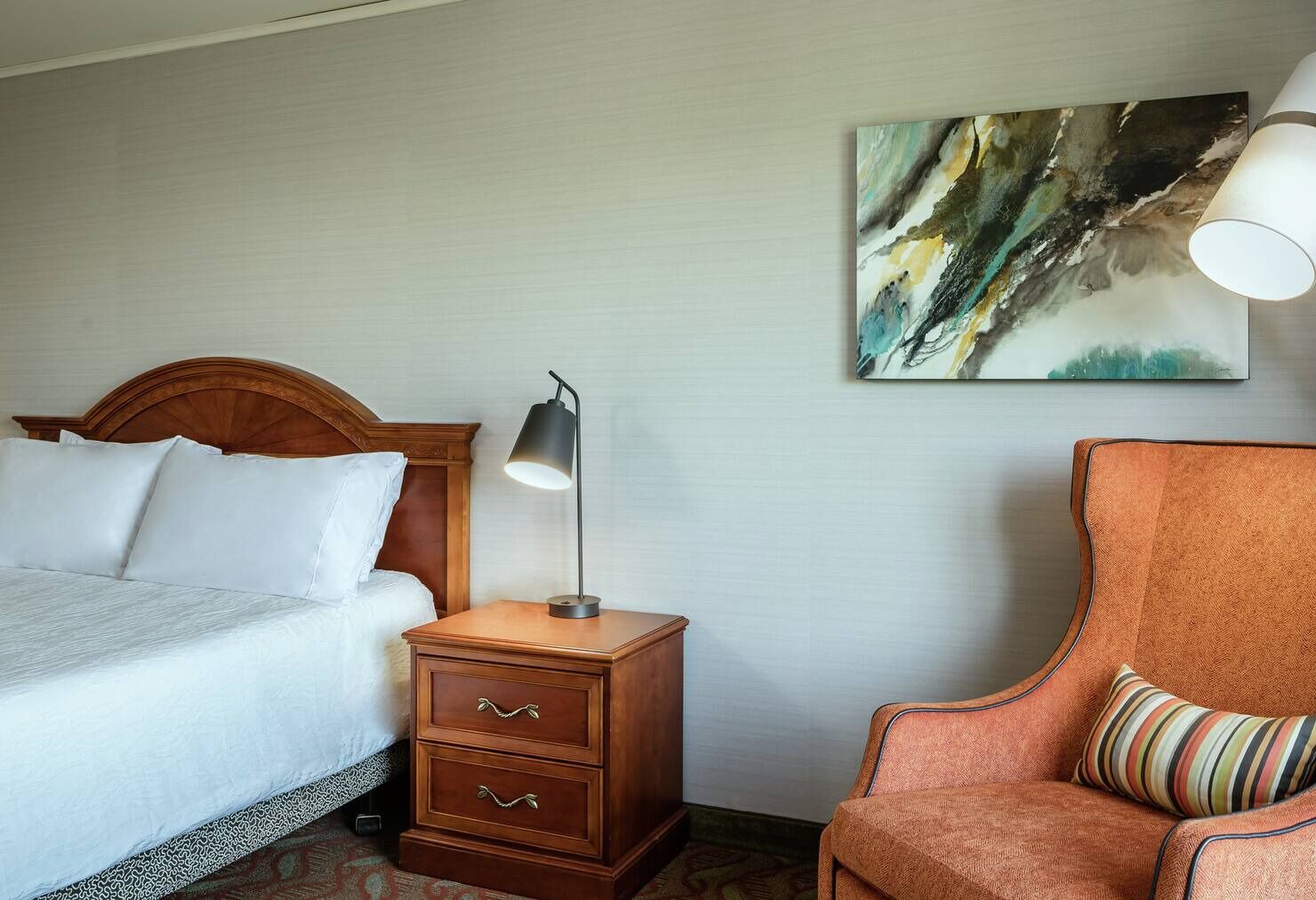 A hotel room with a bed, a nightstand with a lamp, a chair, and a painting on the wall.