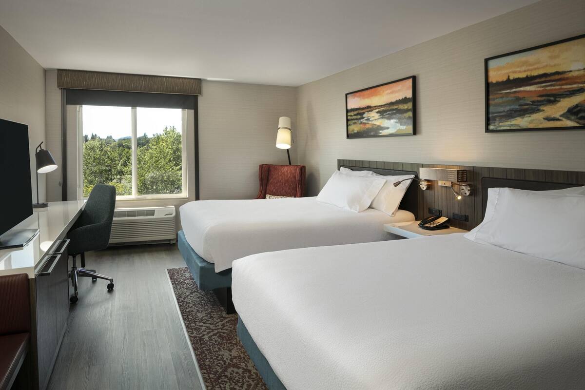 Een hotelkamer met twee bedden, een bureau met stoel, een televisie en een raam met uitzicht op bomen.