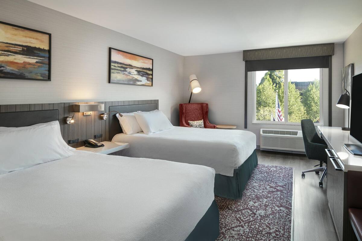 Een hotelkamer met twee bedden, een bureau met stoel, een lamp en een groot raam met een Amerikaanse vlag.