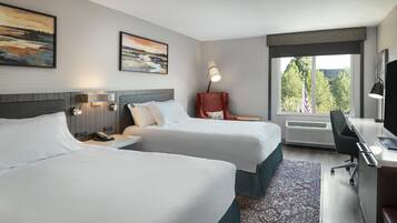 A hotel room with two beds, a desk with a chair, a lamp, and a large window with an American flag.