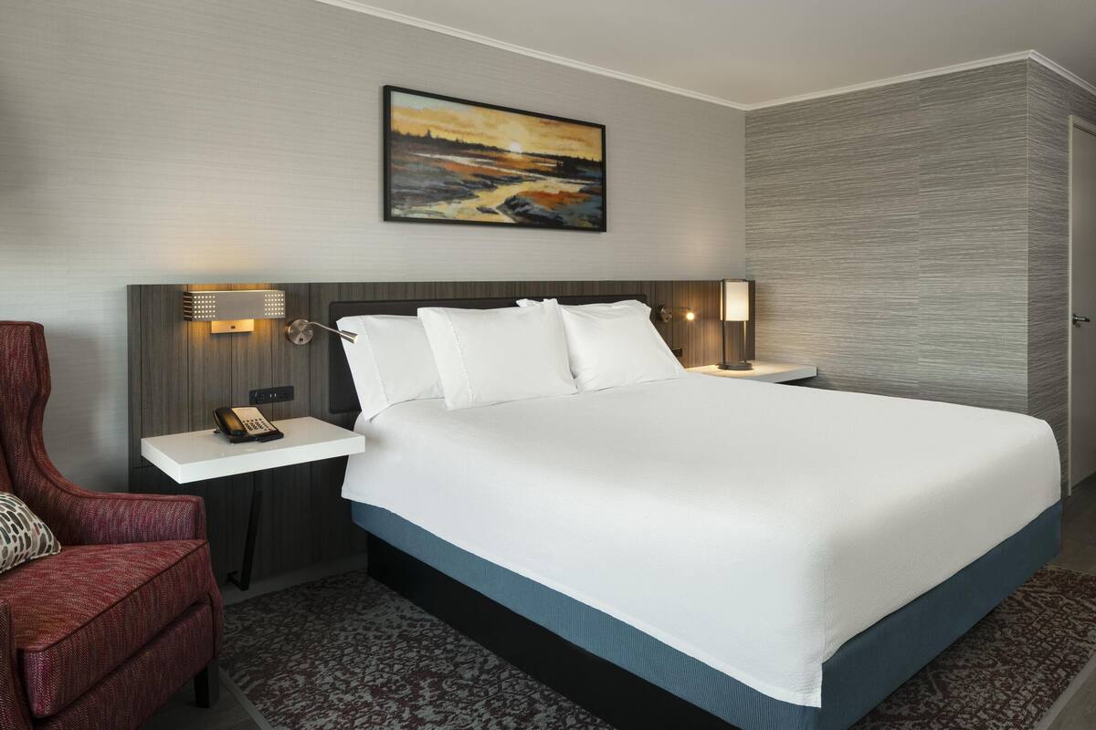 Een hotelkamer met een groot bed, een nachtkastje met een lamp, een stoel en een schilderij aan de muur.