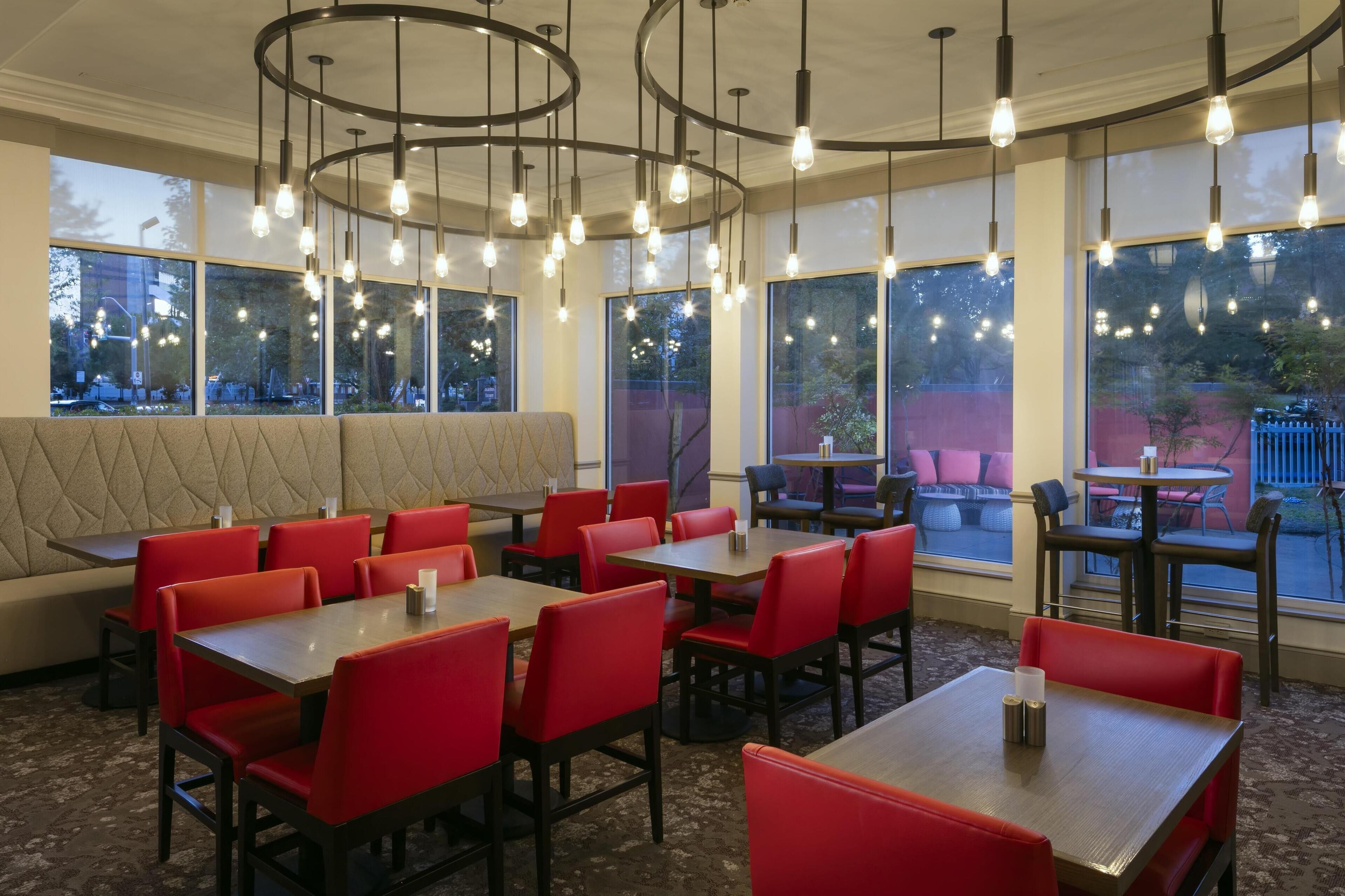 Un restaurant avec des chaises et des tables rouges, de grandes fenêtres et des luminaires suspendus.