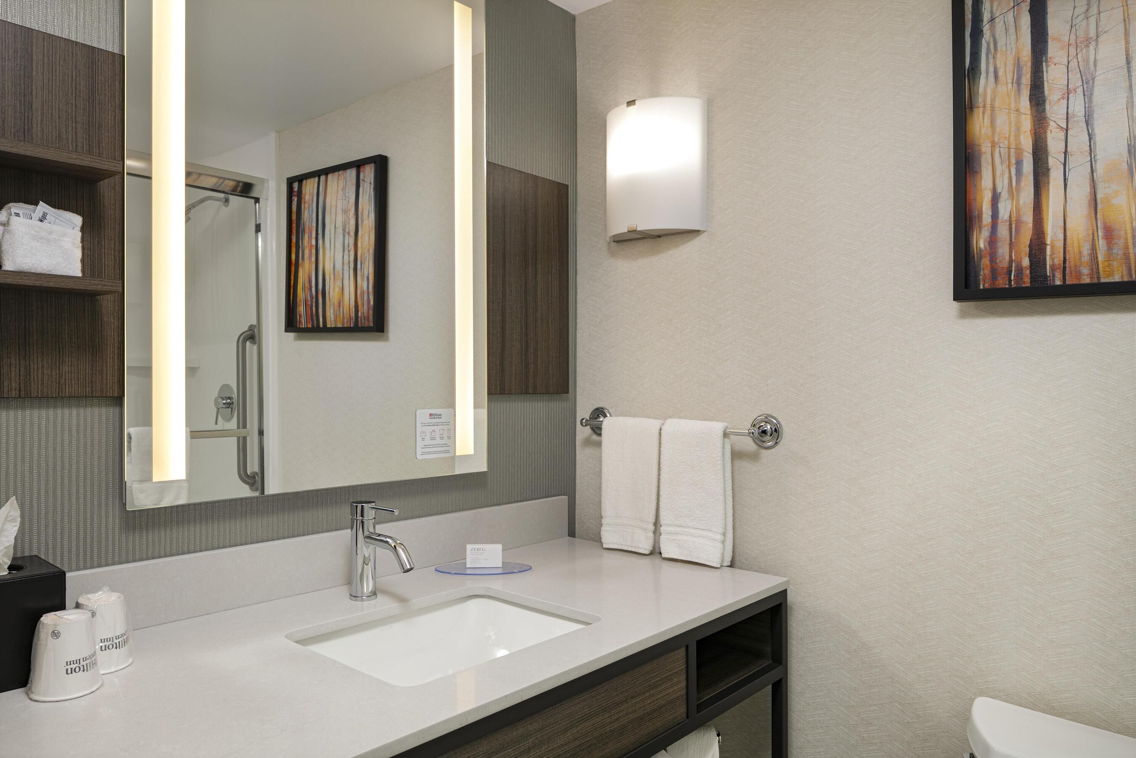 A modern bathroom with a large mirror, a sink, a towel rack, and a framed painting on the wall.